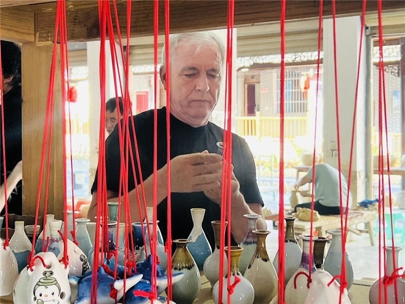 鄉(xiāng)愁如同一根根紅線(xiàn)，將游子與故鄉(xiāng)永遠(yuǎn)地牽連到一起?；逄账囄幕瘎?chuàng)意一條街的母氏陶藝館內(nèi)，紅線(xiàn)吊著陶瓶，思緒游走萬(wàn)里。摩爾多瓦“信息標(biāo)簽”通訊社社長(zhǎng)亞歷山大·塔納斯端詳著館內(nèi)展示的陶藝作品，并挑選出心儀的小擺件帶回摩爾多瓦，也帶走花茂村的“鄉(xiāng)愁”。（張亞欣攝）